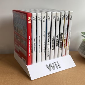 May include: A white plastic display stand holding ten Nintendo Wii game cases in a row. The stand features the Wii logo on the front. The game titles include New Super Mario Bros. Wii, Super Mario Galaxy, Super Smash Bros. Brawl, The Legend of Zelda: Twilight Princess, Rock Band 3, Rock Band 2, and Guitar Hero 5.