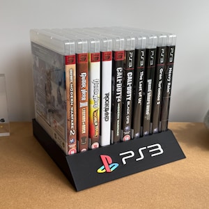 May include: A black plastic display stand holding a row of PlayStation 3 video game cases. The stand features the PS3 logo in blue, red, green, and yellow on the front. The game titles include Call of Duty, Guitar Hero, and The Last of Us.