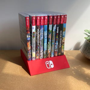 Op de afbeelding: Een rode plastic standaard met tien verticaal geplaatste Nintendo Switch-gamehoesjes. De standaard heeft een wit Nintendo Switch-logo aan de voorzijde. Links staat een doorzichtig acrylblok met de zwarte tekst DISCONEXT MODS erop gedrukt.