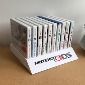 May include: A white plastic display stand holding a row of ten Nintendo 3DS game cases. The stand features the black and red Nintendo 3DS logo on the front angled panel. The game cases are arranged vertically, showing their white spines with titles.