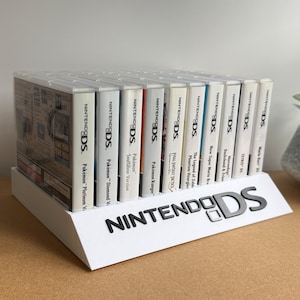 May include: A white plastic display stand holding a row of eleven Nintendo DS game cases. The stand features the black Nintendo DS logo on the front angled panel. The game titles on the spines include various Pokémon editions and other titles.