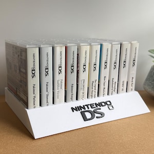 May include: A white plastic display stand featuring the black Nintendo DS logo on the front, holding a row of eleven Nintendo DS game cases. The spines of the games are visible, including titles like Pokémon Platinum, Diamond, SoulSilver, and Ranger.