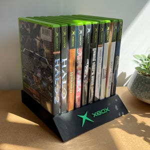 May include: A black angled display stand featuring a green Xbox logo, holding a row of nine video game cases. The cases include titles such as Halo 2, Halo, Morrowind, Fable, Splinter Cell, Grand Theft Auto, Need for Speed, and Soulcalibur II.