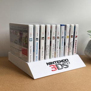 May include: A white plastic display stand featuring the Nintendo 3DS logo in black and red text, holding a row of ten Nintendo 3DS game cases. The stand is angled to showcase the game spines, which include titles like Pokemon, Super Mario, and Zelda.