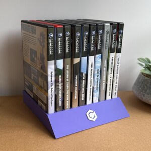 May include: A purple plastic display stand holding ten Nintendo GameCube video game cases in a row. The stand features a small white logo on the front. To the left sits a clear acrylic block with the black text DISCONEXT MODS printed on it.