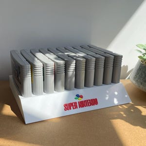 Puede incluir: Un soporte de exhibición blanco que sostiene nueve cartuchos de juego Super Nintendo grises. El soporte presenta el texto rojo "SUPER NINTENDO" y un logotipo colorido. A la izquierda, un letrero acrílico transparente muestra el logo y texto negro "DISCONEXT MODS".