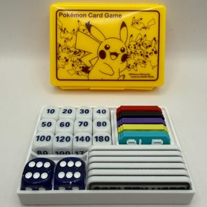 Könnte beinhalten: Ein gelbes Pokémon Card Game Aufbewahrungsetui mit einer Pikachu-Illustration, dazu ein weißes Fach mit weißen Schadensmarken-Würfeln mit schwarzen Zahlen, zwei dunkelblauen Würfeln, mehreren farbigen Kunststoffmarkern und weißen Spielsteinen.