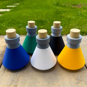 Puede incluir: Una colección de cinco botellas cónicas de colores con cuellos gris claro y tapones de madera. Las botellas son azules, verdes, blancas, negras y amarillas. Están dispuestas sobre una superficie de piedra con un fondo de hierba verde.