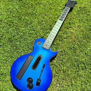 May include: A blue electric guitar-style video game controller featuring a black neck with five fret buttons, a black bridge, a whammy bar, and a strum bar. The headstock displays the Gibson logo in white text against a black background.