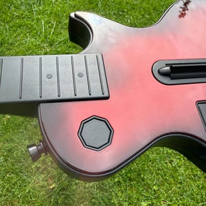 May include: A red and black sunburst guitar controller featuring a black neck with fret markers, a central black slot, and a black octagonal button on the body. The instrument has a glossy finish and a metallic strap button on the lower side.