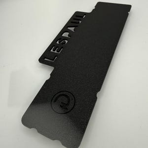 Puede incluir: Una pieza de guitarra de metal negro con la palabra "LESPAUL" recortada. La pieza tiene una superficie texturizada y un logotipo circular. La forma es rectangular con un extremo curvo y varios recortes.