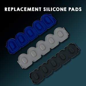 Könnte beinhalten: Drei Ersatz-Silikonpads in Blau, Grau und Schwarz. Jedes Pad hat eine Reihe von ovalen Vertiefungen. Der Text "REPLACEMENT SILICONE PADS" befindet sich oben im Bild.