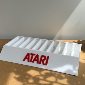 May include: A white plastic display stand featuring ten vertical slots for organizing items. The front of the stand displays the red Atari logo in raised lettering. The structure has a slanted front face and a rectangular base for stability.
