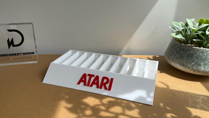 May include: A white plastic display stand featuring ten vertical slots for organizing items. The front of the stand displays the red Atari logo in raised lettering. The structure has a slanted front face and a rectangular base for stability.