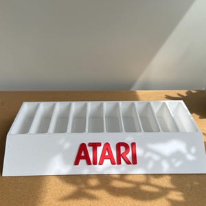 May include: A white plastic storage organizer featuring ten vertical slots for game cartridges. The front of the organizer displays the red Atari logo in raised lettering. The unit has a slanted front face and a rectangular base for stability.