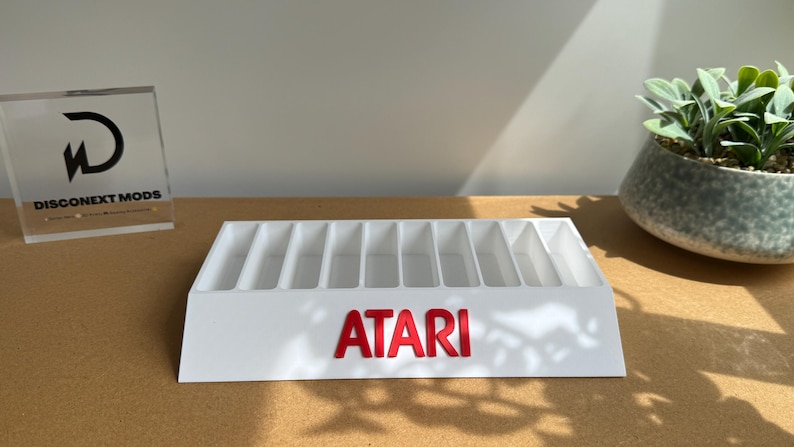 May include: A white plastic storage organizer featuring ten vertical slots for game cartridges. The front of the organizer displays the red Atari logo in raised lettering. The unit has a slanted front face and a rectangular base for stability.