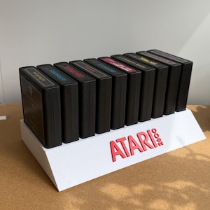 May include: A white display stand with the red text ATARI 2600 holds nine black rectangular game cartridges in a row. To the left, a clear acrylic block features the black logo and text DISCONEXT MODS. The cartridges have colored labels.