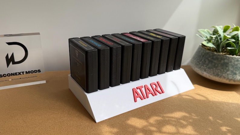 May include: A white plastic display stand featuring the red text ATARI, holding a row of nine black rectangular video game cartridges. The stand is angled to showcase the labels of the cartridges, which are arranged side-by-side in a single line.