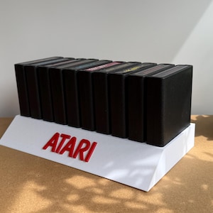 May include: A white display stand featuring the red Atari logo, holding a row of nine black rectangular video game cartridges. The stand is angled, and the cartridges are arranged vertically in a single line, showcasing their black plastic casings.
