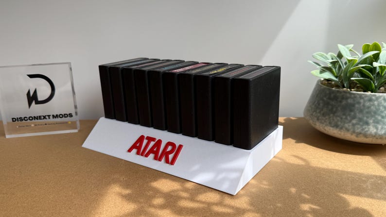 May include: A white display stand featuring the red Atari logo, holding a row of nine black rectangular video game cartridges. The stand is angled, and the cartridges are arranged vertically in a single line, showcasing their black plastic casings.