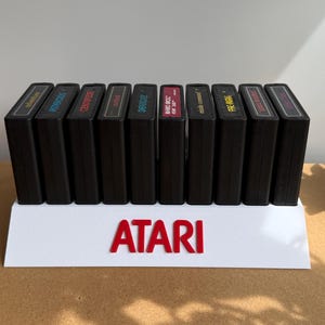 May include: A white plastic display stand featuring the red Atari logo, holding ten black rectangular video game cartridges in a row. The cartridges have various colored labels on their top edges. A clear acrylic sign with the text DISCONEXT MODS is visible to the left of the stand.