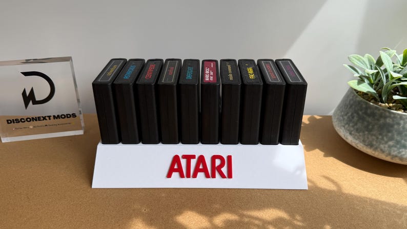 May include: A white plastic display stand featuring the red Atari logo, holding ten black rectangular video game cartridges in a row. The cartridges have various colored labels on their top edges. A clear acrylic sign with the text DISCONEXT MODS is visible to the left of the stand.