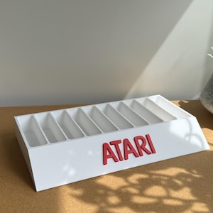 May include: A white rectangular storage stand featuring nine vertical slots for organizing items. The front face displays the word ATARI in bold red capital letters. The structure is angled, providing a clear view of the interior compartments.