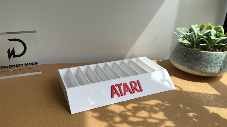 May include: A white rectangular storage stand featuring nine vertical slots for organizing items. The front face displays the word ATARI in bold red capital letters. The structure is angled, providing a clear view of the interior compartments.
