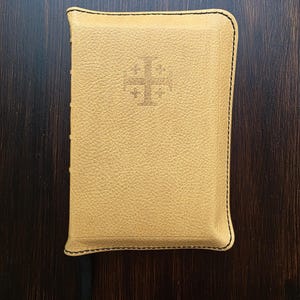 May include: A tan leather-bound book with a cross symbol embossed on the cover. The book has black stitching around the edges and a textured surface. The background is a dark wood grain.