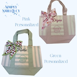 May include: Two canvas tote bags featuring vertical stripes, one in pink and one in green. Each bag displays the Trader Joe's logo, a personalised name, a decorative fabric bow, and a small gold-toned charm with three dangling white pearls.