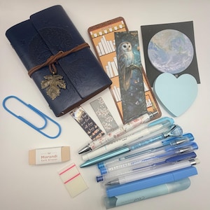 May include: A dark blue leather-bound journal with a brown cord and leaf charm, a large blue paperclip, a Morandi soft eraser, two small adhesive tabs, three bookmarks, a heart-shaped sticky note, a round earth sticky note, and several blue pens.