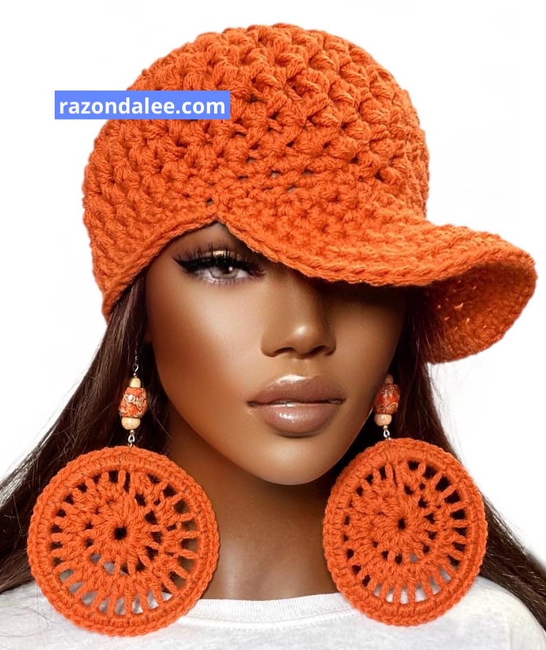 May include: An orange crocheted baseball cap with a textured knit pattern and the text razondalee.com. Large, round, orange crocheted earrings with a circular openwork design and small wooden beads hang from the ears.