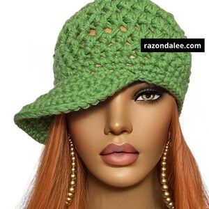 May include: A green crocheted cap with a visor. The cap is made of a textured, open-weave yarn. The text "razondalee.com" is displayed on the cap. The cap is worn by a mannequin with long, orange hair and gold earrings.