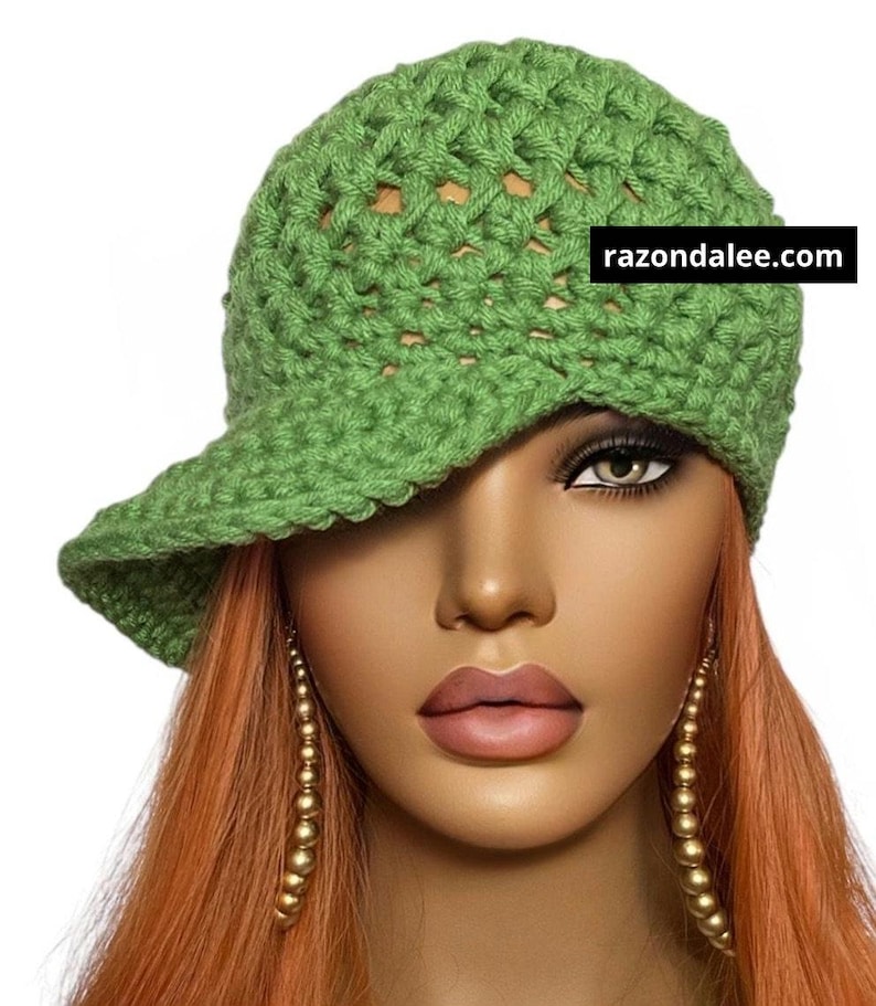 May include: A green crocheted cap with a visor. The cap is made of a textured, open-weave yarn. The text "razondalee.com" is displayed on the cap. The cap is worn by a mannequin with long, orange hair and gold earrings.