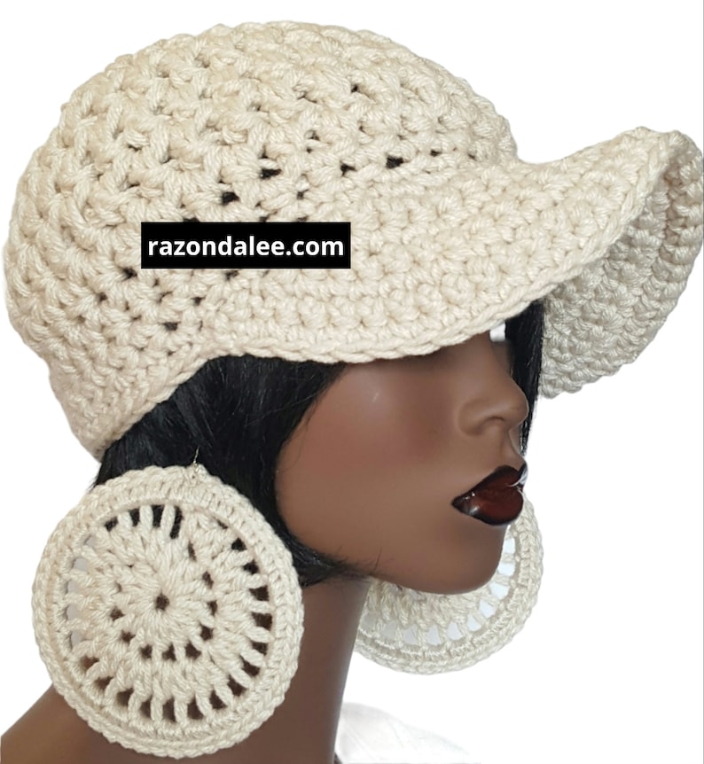 May include: A cream-coloured crocheted cap with a wide brim and a black rectangular label featuring the white text razondalee.com. The cap is paired with large, circular, cream-coloured crocheted earrings with a radial pattern.