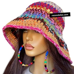May include: A multi-colored crochet bucket hat featuring horizontal stripes in shades of red, orange, yellow, green, blue, and purple, finished with a bright pink braided edge. The text razondalee.com is visible on the side of the hat.