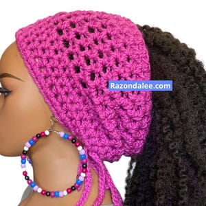 May include: A bright pink crocheted hair accessory with a textured, open-stitch pattern and dangling ties. The accessory is worn on a mannequin head with dark, curly hair and large hoop earrings featuring multicolored beads. Text reads Razondalee.com.