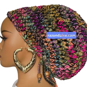 May include: A crochet beanie featuring a variegated yarn pattern in shades of hot pink, black, olive green, and orange. The hat includes two dangling cords with wooden beads at the ends and a blue rectangular label with the text razondalee.com.