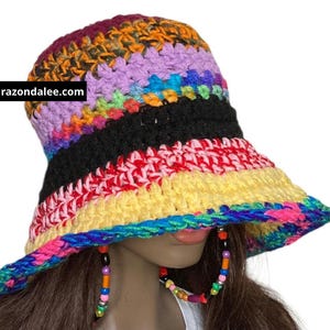 May include: A multi-colored crochet bucket hat featuring horizontal stripes in shades of purple, orange, black, red, yellow, and blue. The hat includes a black rectangular tag with the white text razondalee.com and two beaded chin straps.