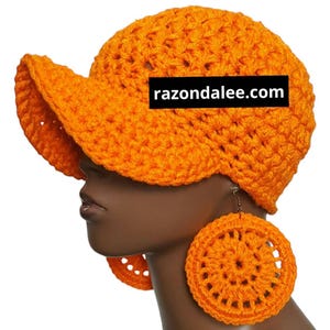 May include: Bright orange crochet hat with a visor and matching circular earrings. The hat and earrings are made of a thick, textured yarn. The website razondalee.com is printed on the hat.
