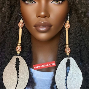 May include: A pair of long dangle earrings featuring large, flat, silver-toned cowrie shell-shaped pendants. Each earring includes a vertical arrangement of two brown and white zebra-striped beads, a small gold-toned bead, and a bamboo tube. Text reads razondalee.com.
