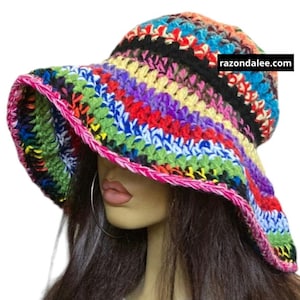 May include: A multi-colored crochet bucket hat featuring horizontal stripes in vibrant shades of red, blue, green, yellow, purple, and black. The wide brim is edged with a bright pink trim. The text razondalee.com is printed on the hat.