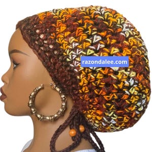 May include: A crochet beanie in a variegated yarn featuring shades of brown, orange, yellow, and white. The hat has a textured stitch pattern and includes dangling brown braided ties with two orange beads. A blue label displays razondalee.com.