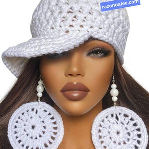 May include: A white crocheted cap with a brim and matching large circular crocheted earrings featuring white beads. The cap displays the text razondalee.com on a blue rectangular background. The design is consistent in colour and texture.