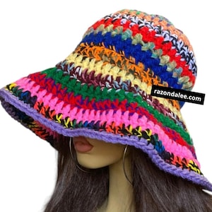May include: A colorful, handmade crochet bucket hat. The hat features a wide brim and a rounded crown, with a vibrant mix of pink, red, orange, yellow, green, blue, and purple yarn. The brim is trimmed with a purple border. The text "razondalee.com" is visible on the hat.