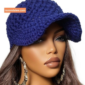 May include: A deep blue crochet baseball cap featuring a textured, woven pattern and a curved brim. The cap includes an orange rectangular label with the white text razondalee.com positioned on the upper left side of the crown.