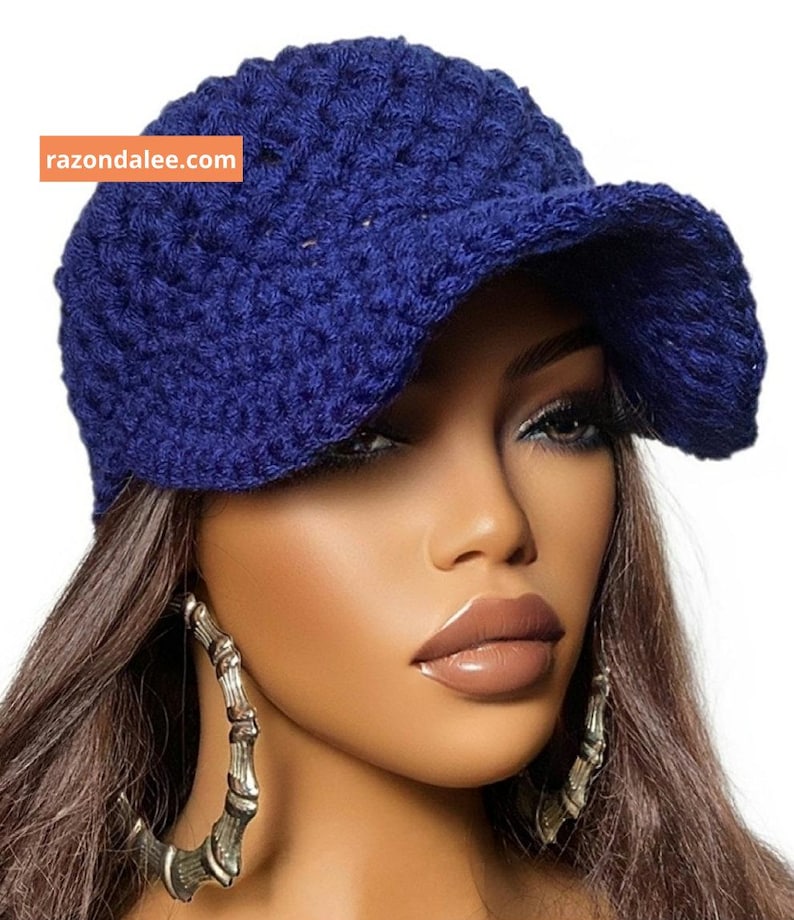 May include: A deep blue crochet baseball cap featuring a textured, woven pattern and a curved brim. The cap includes an orange rectangular label with the white text razondalee.com positioned on the upper left side of the crown.
