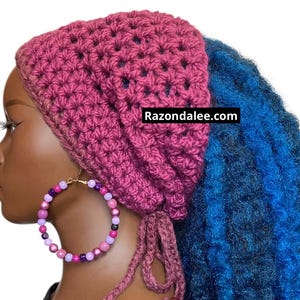 May include: A dark magenta crocheted head covering with a textured star-like stitch pattern and two dangling ties at the base. The accessory features the text Razondalee.com and is paired with a large hoop earring made of purple, pink, and black beads.