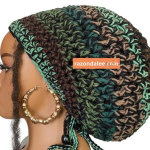 May include: A crochet beanie featuring a camouflage-inspired pattern in shades of black, dark green, light green, and brown. The hat includes a drawstring with a dark bead and displays the text razondalee.com on an orange rectangular label.