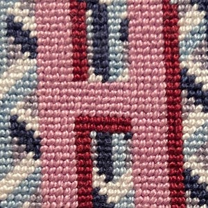 May include: Close-up of a needlepoint design featuring the letter "H". The letter is a light pink color with a dark red outline. The background is a mix of blue, white, and light blue stitches.